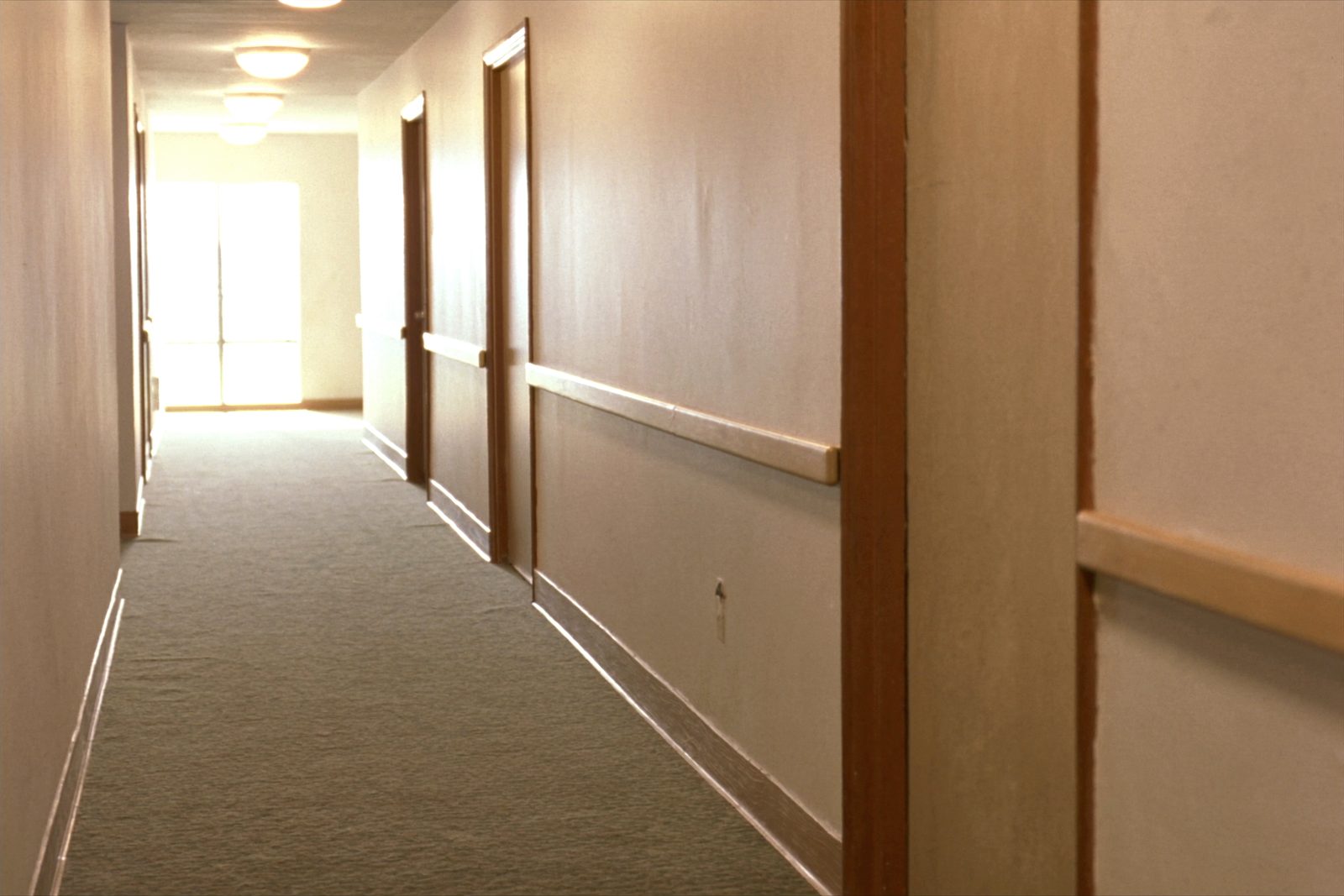 A daylit corridor in a small senior apartment building, with a handrail running toward a bright window at the far end.