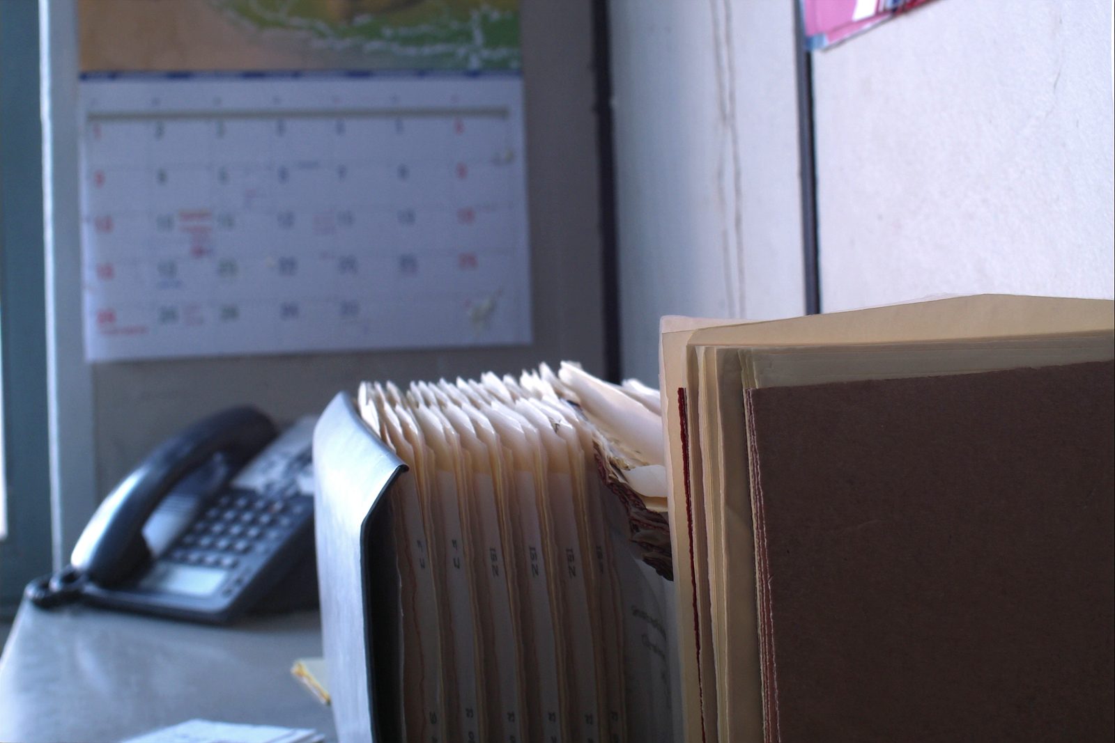 An open accordion file of tabbed dividers and a stack of folders on a desk beside a landline telephone.
