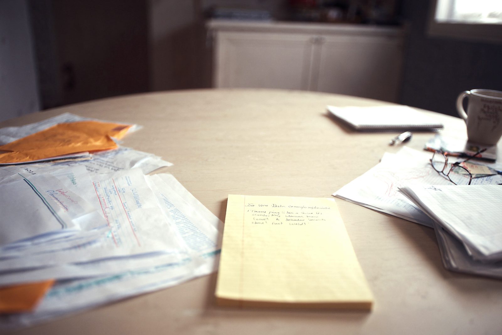 A kitchen table covered with opened plan mail, statements and a handwritten legal pad, with reading glasses and a coffee mug pushed to one side.